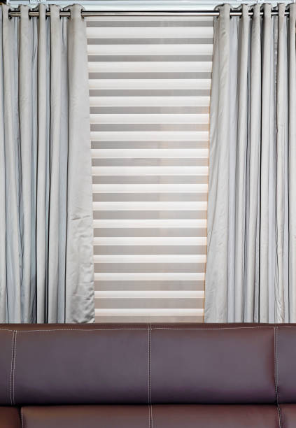 Decorative silver drapes behind brown leather couch wooden blinds